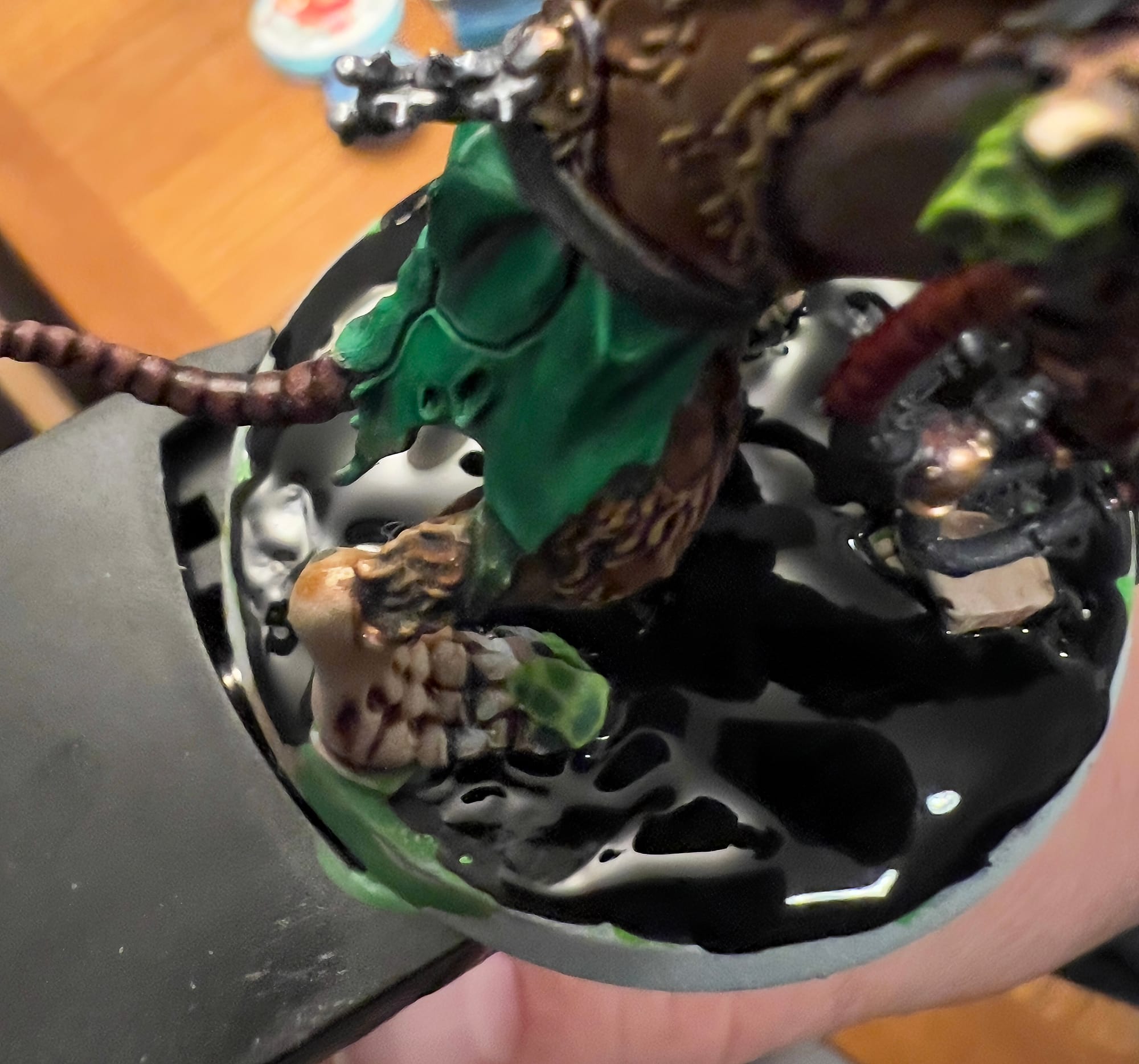Close-up of a miniature figure’s base covered in glossy black liquid texture, with a brown, furred leg and green cloak visible above, suggesting a wet or oily surface effect.