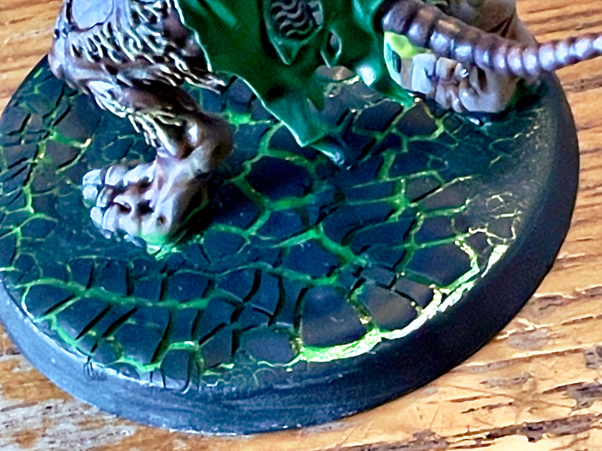 Close-up of a miniature figure’s base showing cracked, textured ground painted dark with glowing green fissures, suggesting eerie, magical energy.