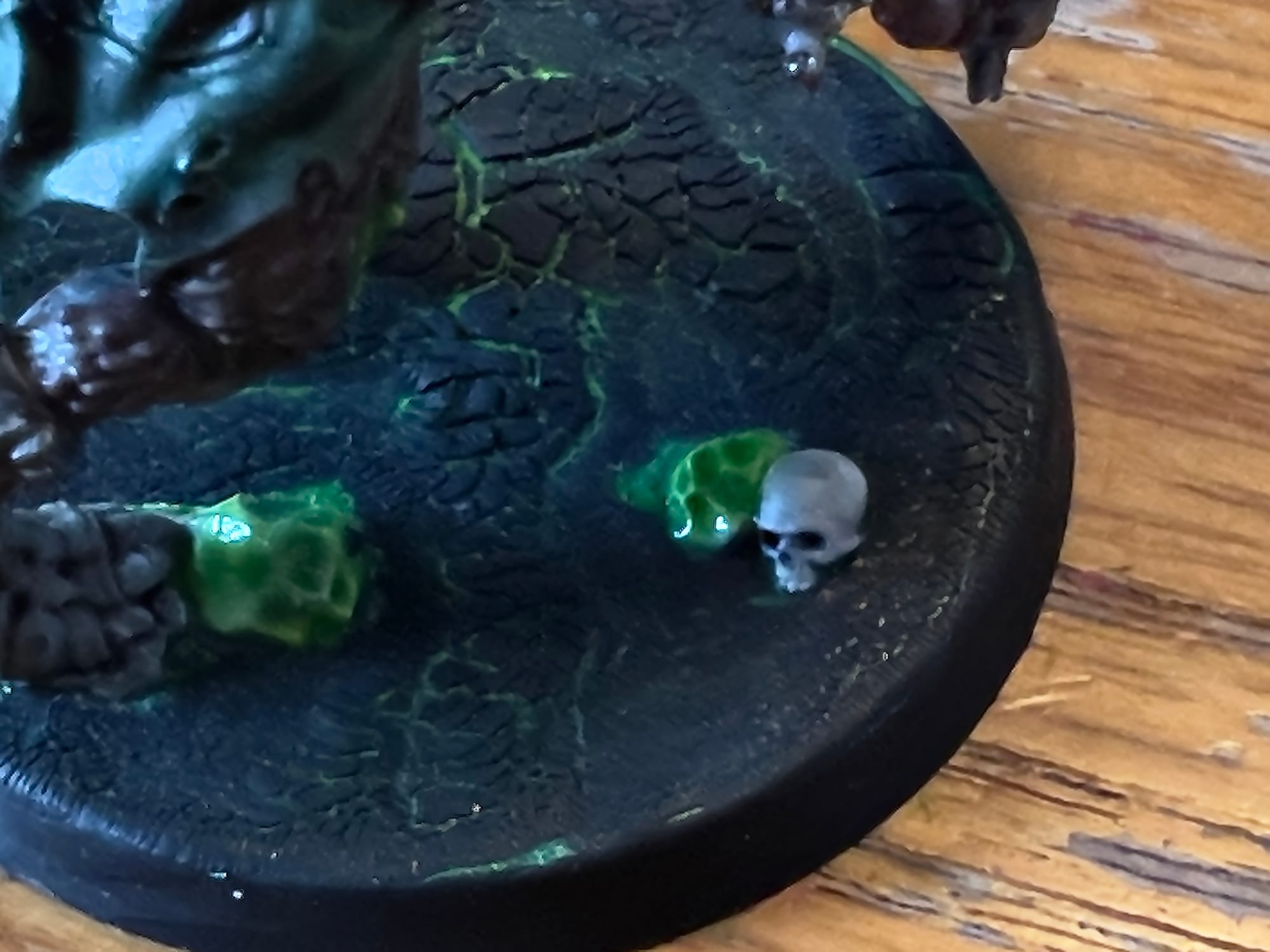 Close-up of a miniature figure’s base painted with cracked dark ground glowing green underneath, featuring two small green slime patches and a grey skull for detail.