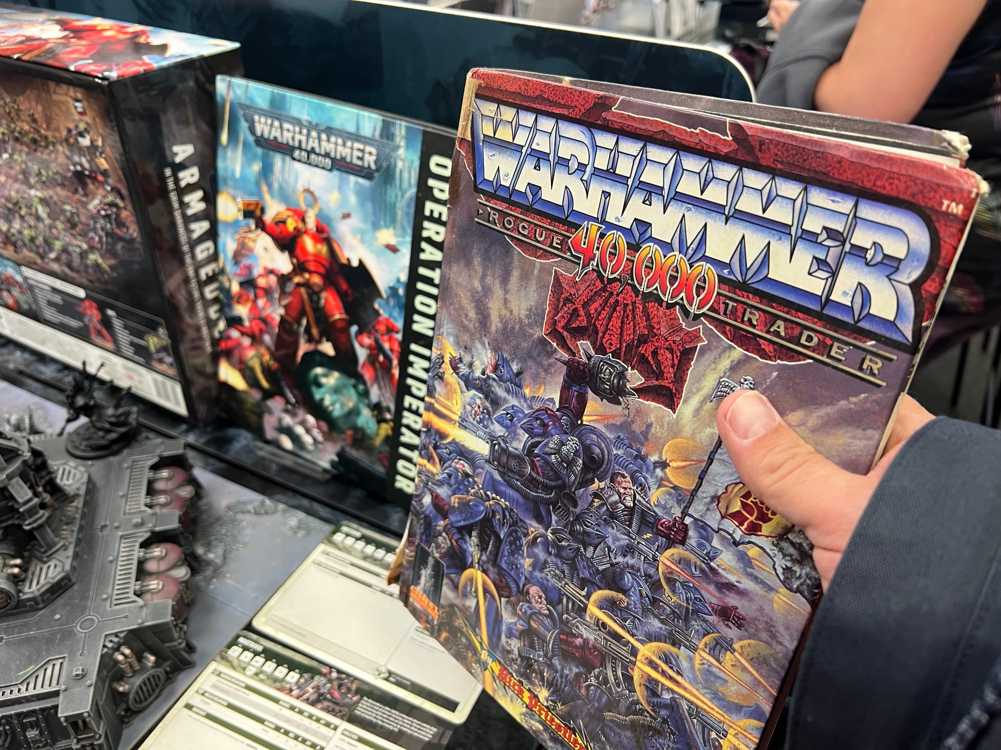 A hand holds a well-worn copy of Warhammer 40,000: Rogue Trader at a gaming display, surrounded by modern 11th edition Warhammer books, miniatures and boxed sets.