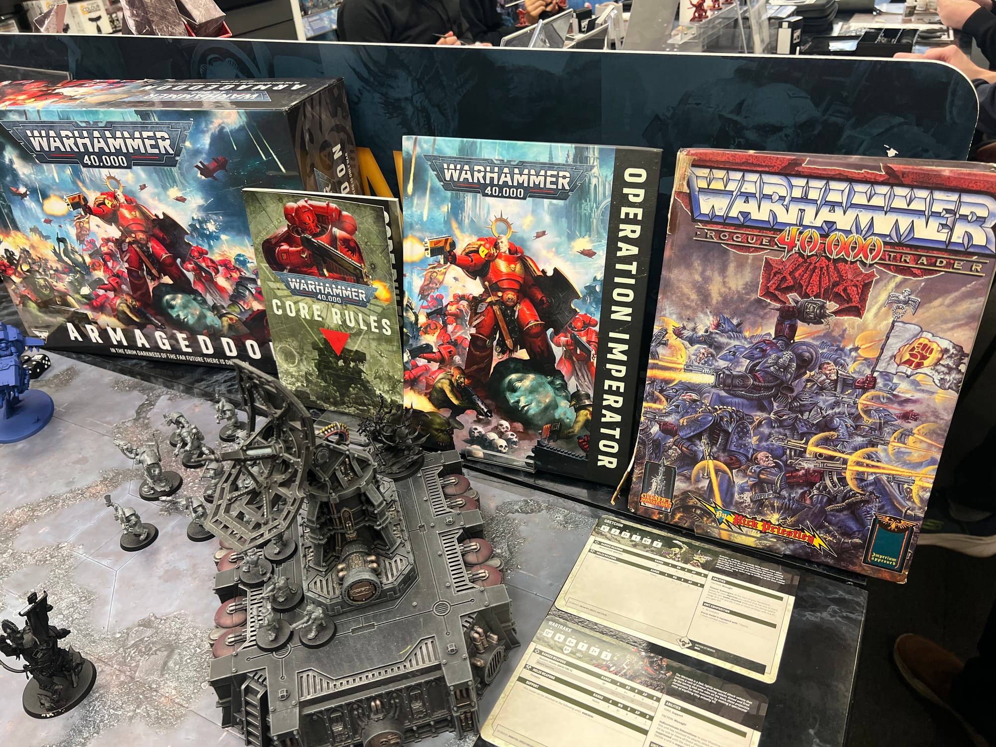 Display of Warhammer 40,000 books and miniatures, featuring the modern Armageddon and Operation Imperator volumes alongside a vintage Warhammer 40,000: Rogue Trader rulebook and painted terrain pieces.