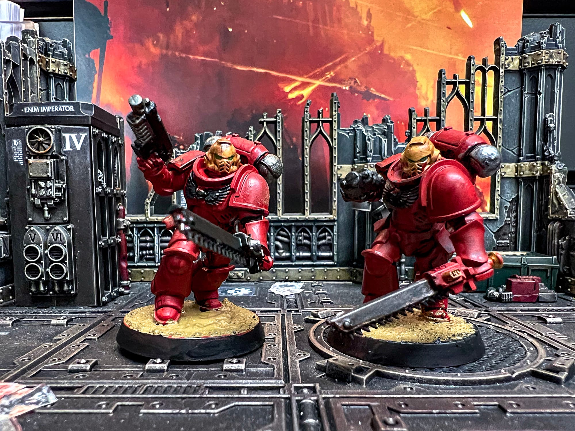 Two red Space Marine miniatures armed with chainswords and pistols advance through ruined gothic scenery, with a fiery battlefield backdrop behind them.